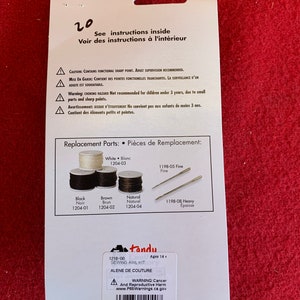 May include: A white and blue packaging for a sewing awl kit. The packaging includes a warning label about cancer and reproductive harm. The text on the packaging reads "Tandy Sewing AWL Kit" and "Alene de Couture". The packaging also includes a list of replacement parts with their corresponding numbers.