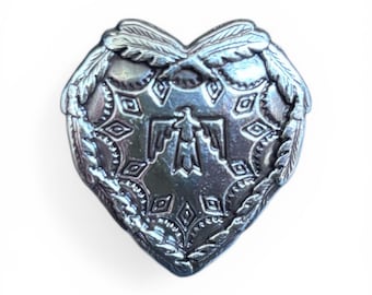 Puede incluir: Un colgante plateado en forma de corazón con un diseño en relieve. El colgante presenta un símbolo central de Thunderbird, rodeado de patrones geométricos y detalles de hojas. El diseño general tiene una estética vintage o del suroeste.