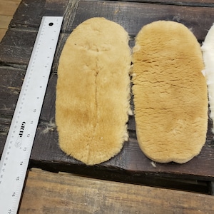 May include: Two tan-colored sheepskin insoles, one slightly larger than the other, are laid out on a wooden surface. A ruler is visible in the background.