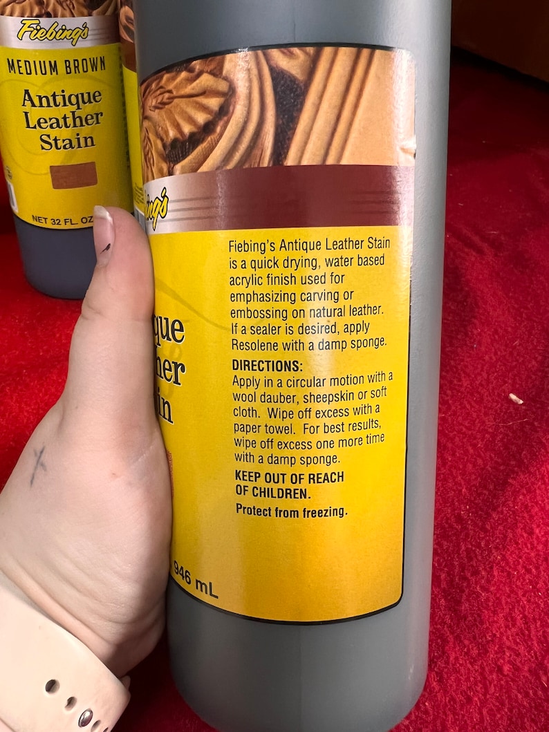 May include: A bottle of Fiebing's Antique Leather Stain in Medium Brown. The bottle is 32 fluid ounces and is labeled with instructions for use. The label is yellow with black text. The bottle is on a red surface.