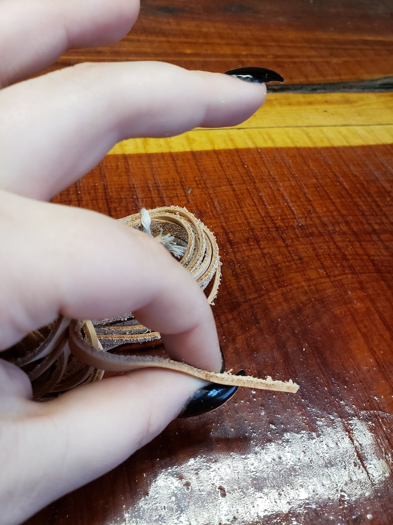 K&ouml;nnte beinhalten: Nahaufnahme einer Hand, die eine Spule mit hellbraunem Lederband h&auml;lt. Das Band ist etwa 6 mm breit und zu einem lockeren Kreis gewickelt. Die Hand h&auml;lt das Band in der Mitte der Spule, und das Band wird aus der Spule gezogen.