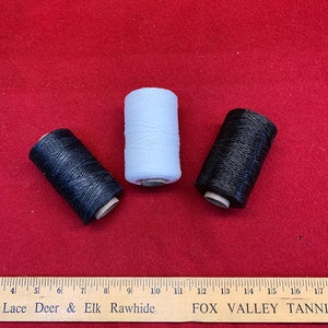 May include: Three spools of waxed thread, one white and two black, on a red surface. The spools are labeled "Lace Deer & Elk Rawhide" and "FOX VALLEY TANNERY".
