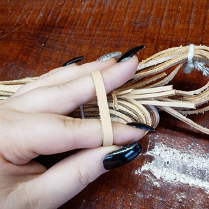 K&ouml;nnte beinhalten: Ein B&uuml;ndel hellbrauner Lederstreifen, zusammengehalten mit einem wei&szlig;en Band, wird in einer Hand mit schwarzem Nagellack gehalten.
