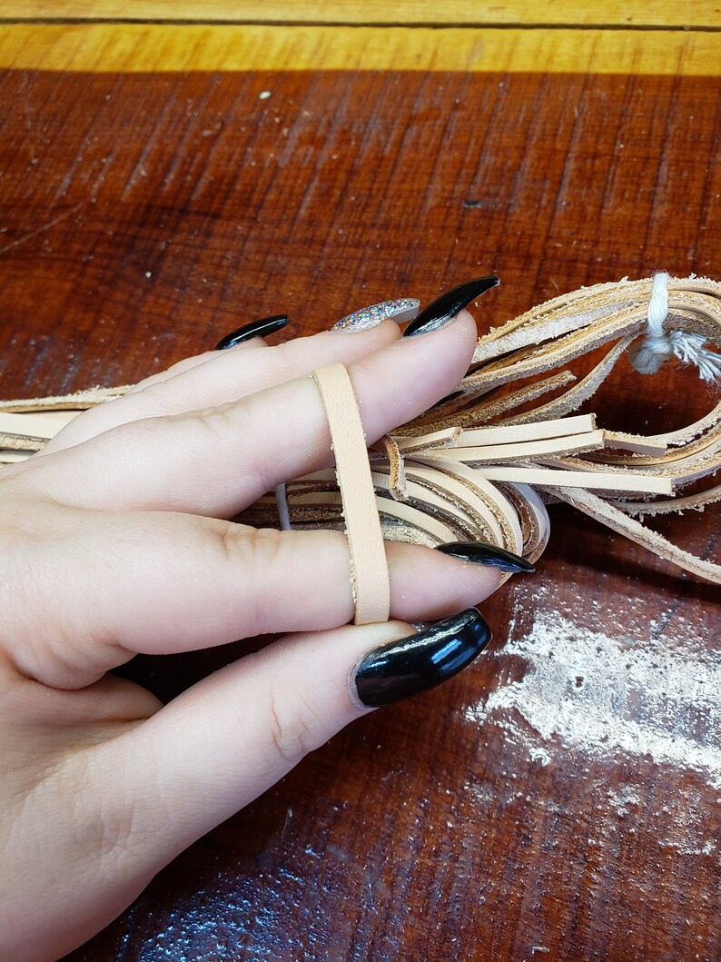 K&ouml;nnte beinhalten: Ein B&uuml;ndel hellbrauner Lederstreifen, zusammengehalten mit einem wei&szlig;en Band, wird in einer Hand mit schwarzem Nagellack gehalten.