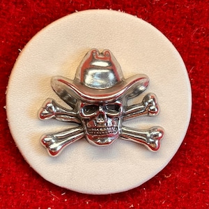 May include: A silver skull wearing a cowboy hat and crossed bones. The skull and bones are on a white background.