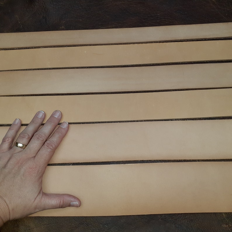 May include: A close-up of six strips of light brown leather laid out on a dark brown surface. The leather is smooth and appears to be of good quality.