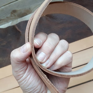 May include: A close-up of a light brown leather strap, folded in half and held in a hand. The strap is smooth and appears to be made of high-quality leather.
