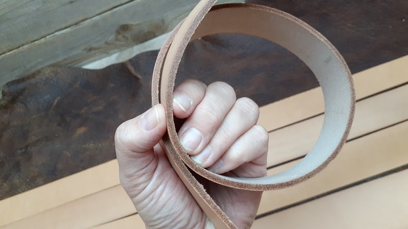 May include: A close-up of a light brown leather strap, folded in half and held in a hand. The strap is smooth and appears to be made of high-quality leather.