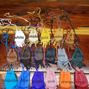 May include: A collection of 16 small leather pouches with fringe, each in a different color. The pouches are arranged in two rows, with the colors labeled below each pouch. The colors include white, smoke, gold, saddle, tobacco, whiskey, chocolate, dark brown, black, grey, green, turquoise, blue, purple, pink, orange, and red.