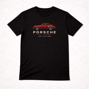 May include: A black t-shirt featuring a graphic of a red Porsche 911 Turbo car. Below the car, the text reads PORSCHE 911 TURBO 930 1975-1989 in white and red lettering. The shirt has short sleeves and a crew neckline.