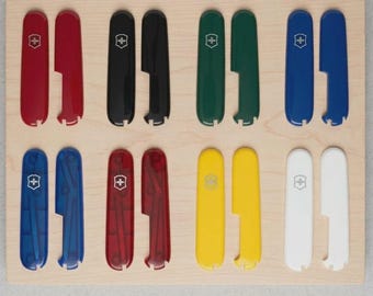 Pode incluir: Um conjunto de doze talas de substituição para canivetes suíços, dispostas em pares. O conjunto inclui talas de plástico sólido nas cores vermelha, preta, verde, amarela e branca, além de opções translúcidas em azul e vermelho, cada uma com o emblema da cruz prateada.