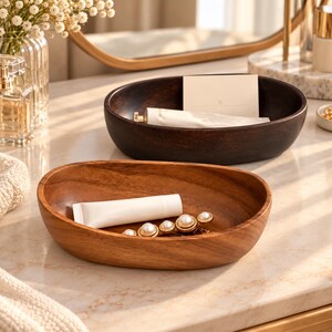 May include: Two oval wooden bowls with smooth, curved edges. The front bowl is light brown and contains a white tube and a pearl-accented gold chain. The back bowl is dark brown and holds a white tube and a rectangular white card.