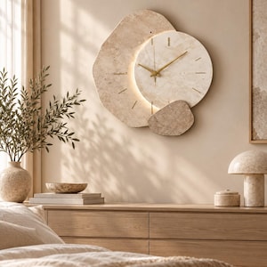 May include: A wall clock featuring an irregular, organic-shaped beige stone base with a circular stone face layered on top. The clock has thin gold-toned hands and gold-toned hour markers, with a soft light glowing from behind the circular face.