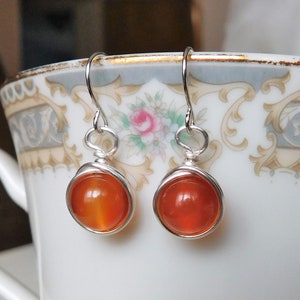 May include: A pair of silver dangle earrings with orange carnelian gemstones. The earrings are hanging from a white porcelain teacup.