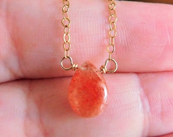 May include: A gold chain necklace with a teardrop-shaped sunstone pendant. The pendant is a light orange color with a shimmery finish.