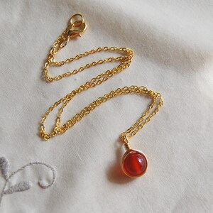 May include: A gold-colored chain necklace with a round, red gemstone pendant. The pendant is wire-wrapped in gold. The necklace is displayed on a white, textured surface.