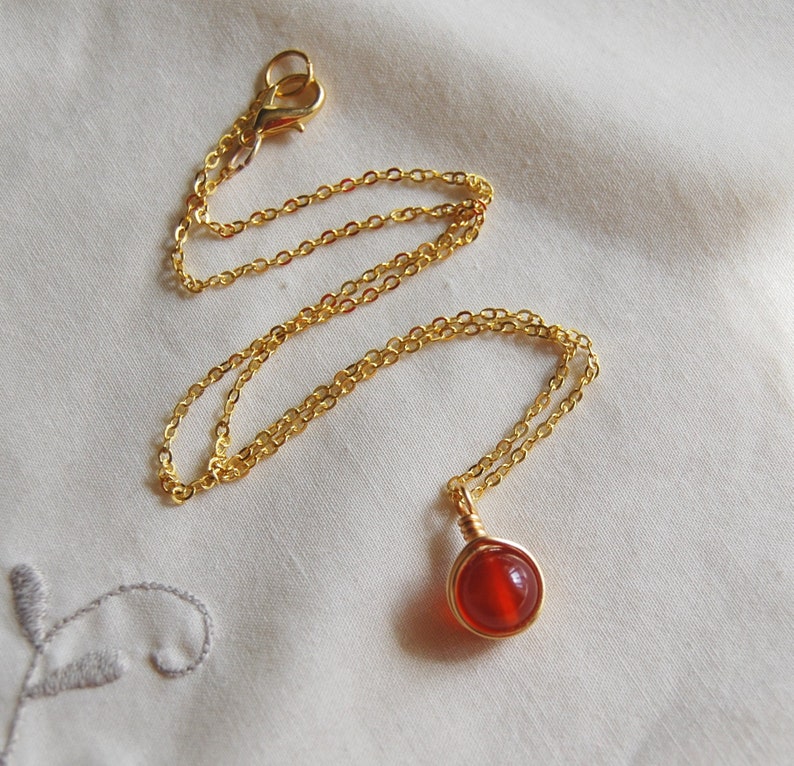 May include: A gold-colored chain necklace with a round, red gemstone pendant. The pendant is wire-wrapped in gold. The necklace is displayed on a white, textured surface.