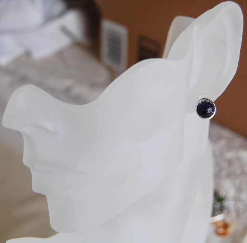 May include: A close-up of a silver stud earring with a dark blue gemstone. The earring is displayed on a white mannequin head.