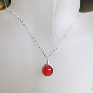 May include: A delicate silver necklace featuring a vibrant red, round gemstone pendant. The gemstone is encased in a wire wrap, suspended from a fine chain. The necklace is displayed on a white mannequin.