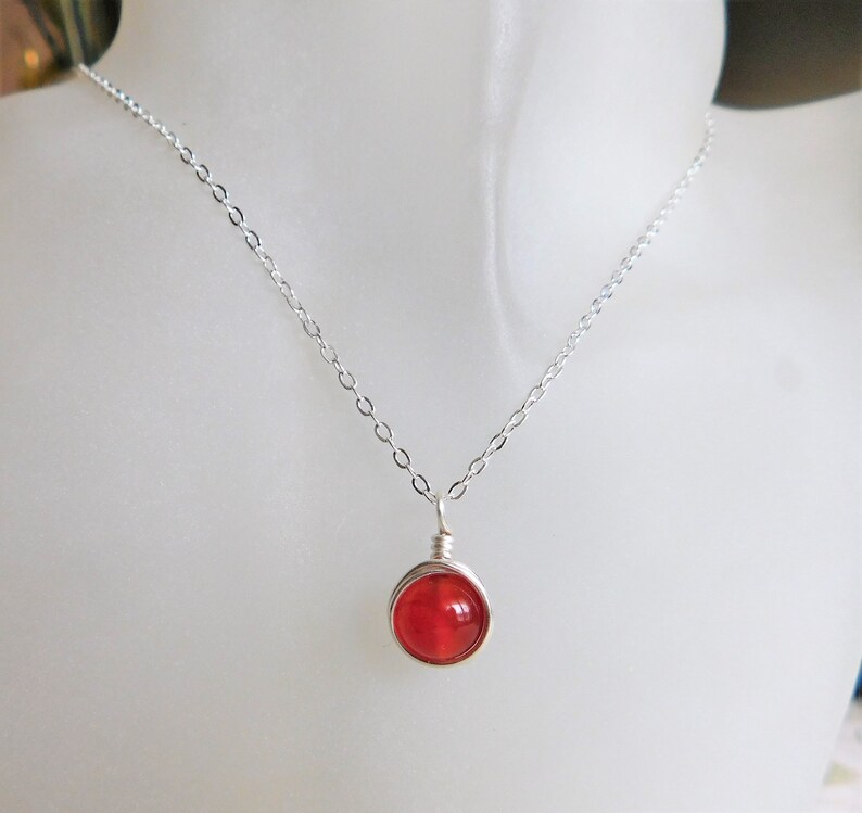 May include: A delicate silver necklace featuring a vibrant red, round gemstone pendant. The gemstone is encased in a wire wrap, suspended from a fine chain. The necklace is displayed on a white mannequin.