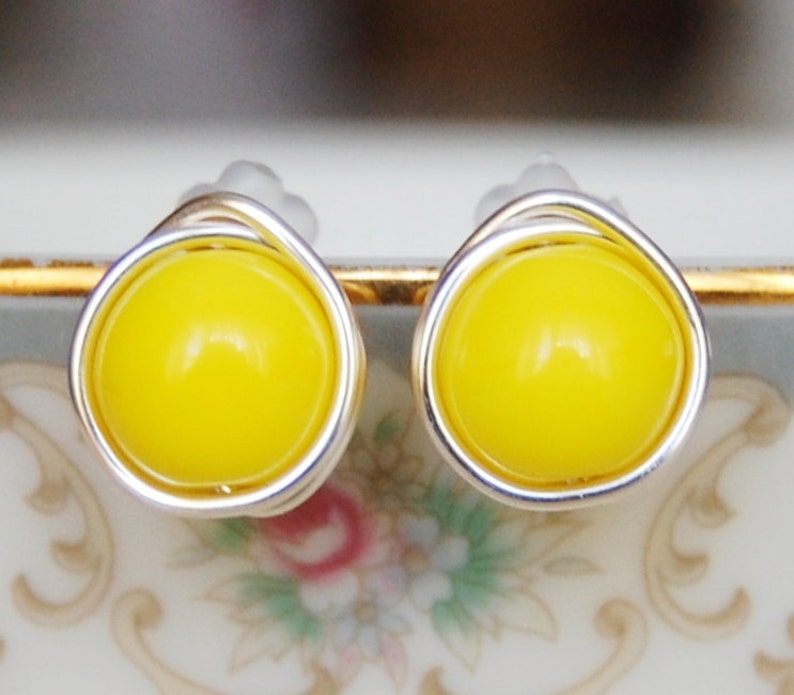 May include: A pair of yellow stud earrings with silver wire wrapping. The earrings are on a white surface with a floral pattern.