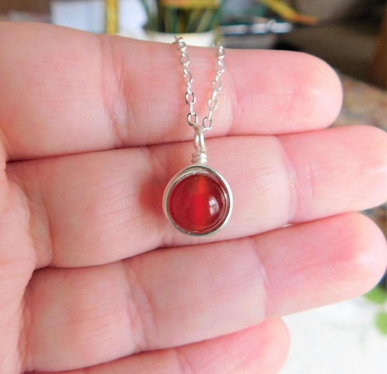 May include: A silver necklace featuring a round, vibrant red gemstone pendant. The gemstone is encased in a silver wire setting and hangs from a delicate silver chain. The pendant is approximately 0.5 inches in diameter.
