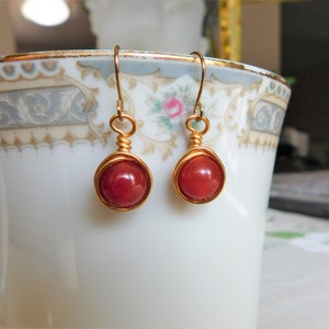 May include: A pair of handmade earrings featuring round, red beads encased in gold-colored wire. The earrings have a simple hook design for pierced ears. The beads are a deep, rich red color, and the wire has a shiny finish.
