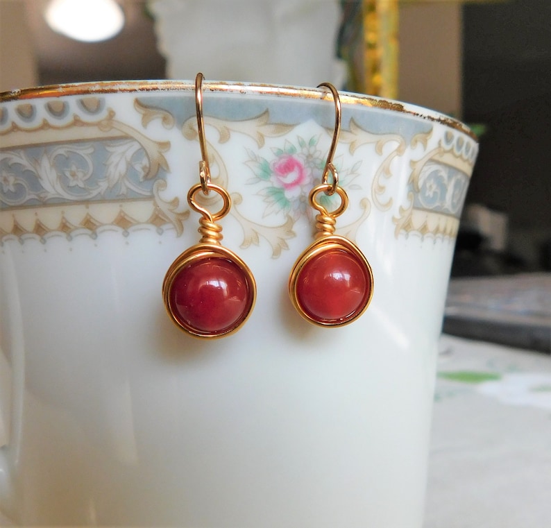 May include: A pair of handmade earrings featuring round, red beads encased in gold-colored wire. The earrings have a simple hook design for pierced ears. The beads are a deep, rich red color, and the wire has a shiny finish.