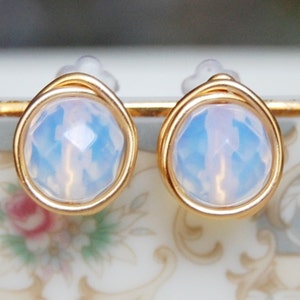 May include: A pair of gold-toned earrings with a white opal stone in each. The stones are faceted and have a light blue hue.