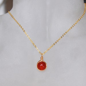 May include: A gold-colored necklace with a small, round, orange gemstone pendant. The gemstone is wrapped in gold wire and hangs from a delicate chain. The necklace is displayed on a neutral background.