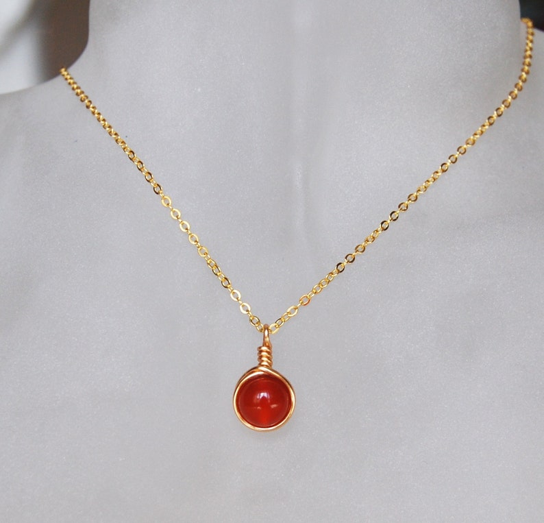 May include: A gold-colored necklace with a small, round, orange gemstone pendant. The gemstone is wrapped in gold wire and hangs from a delicate chain. The necklace is displayed on a neutral background.