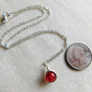 May include: A silver necklace with a small, round, red gemstone pendant. The pendant is wire-wrapped. A U.S. quarter is next to the necklace for size comparison. The chain is delicate and has a lobster clasp.