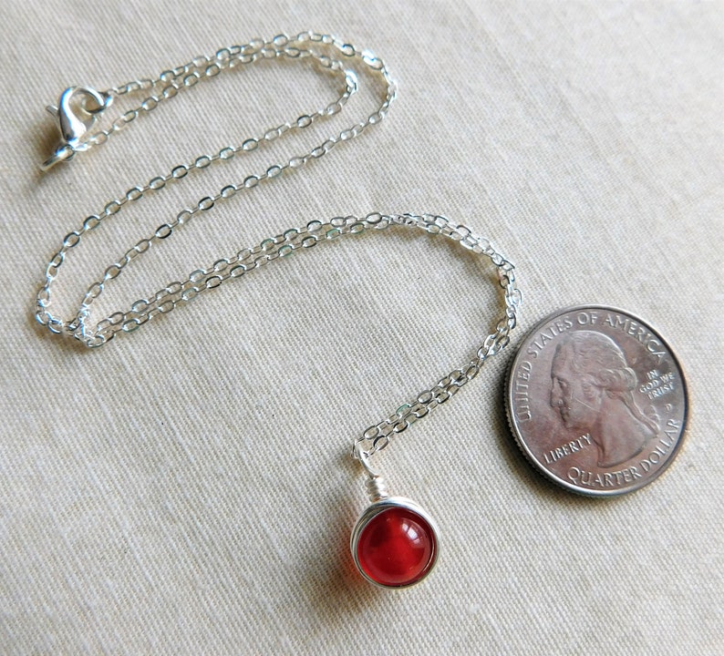 May include: A silver necklace with a small, round, red gemstone pendant. The pendant is wire-wrapped. A U.S. quarter is next to the necklace for size comparison. The chain is delicate and has a lobster clasp.
