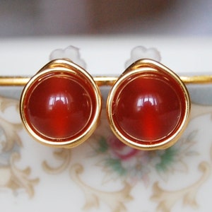 May include: A pair of stud earrings featuring round, polished, reddish-orange gemstones set in gold-colored metal. The earrings have a simple, elegant design, with the gemstone encircled by a thin, twisted wire.