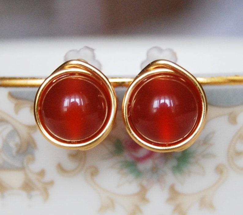 May include: A pair of stud earrings featuring round, polished, reddish-orange gemstones set in gold-colored metal. The earrings have a simple, elegant design, with the gemstone encircled by a thin, twisted wire.