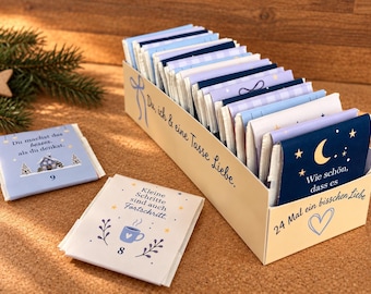 May include: A rectangular cream-colored box containing twenty-four small, individually wrapped packets in shades of blue and white. The box features the text "Du, ich & eine Tasse Liebe" and "24 Mal ein bisschen Liebe" with a heart icon.
