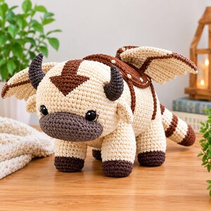 May include: A cream-colored crochet plush toy shaped like a flying bison with dark brown accents on the feet, tail, and a large arrow marking on the head. It features two dark curved horns, black plastic eyes, and a brown saddle on its back.