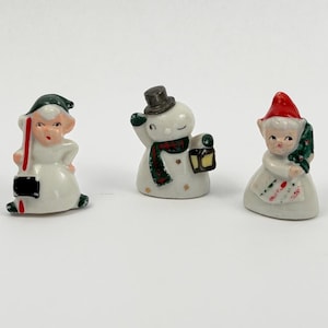 May include: Three small ceramic figurines featuring a white elf in a green hat holding a black shovel, a white snowman wearing a grey top hat and a green patterned scarf holding a lantern, and a white elf in a red hat holding a green pine branch.