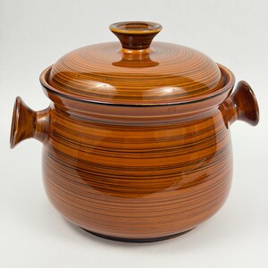 May include: A round, glazed ceramic cooking pot with a matching lid, featuring a glossy finish in shades of brown with thin, horizontal dark brown stripes. The pot includes two side handles and a central knob on the lid.