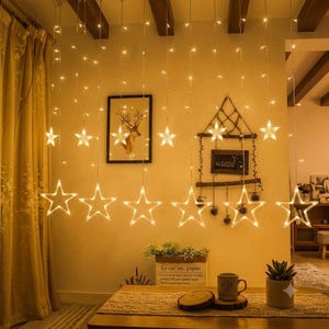 May include: A set of hanging LED string lights featuring multiple illuminated star-shaped ornaments. The warm white lights are arranged in vertical strands, creating a glowing curtain effect against a wall with framed wall art.