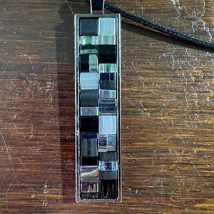 May include: A rectangular pendant necklace featuring a mosaic of small, square, and rectangular tiles in alternating shades of black, silver, and metallic gray, set within a polished silver-toned metal frame on a thin black cord.