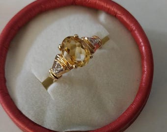 May include: A gold ring with an oval, yellow gemstone as the centerpiece. The ring has small accent stones on either side of the main stone. The ring is presented in a red, circular jewelry box.