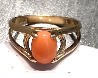 May include: A gold-toned ring featuring an oval, peach-colored gemstone. The ring's band has an open, cage-like design, with the gemstone held securely in the center. The ring is likely made of gold or a gold-colored metal.