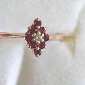 May include: A gold-toned ring featuring a cluster of small, deep red gemstones arranged in a floral pattern. The center of the flower has a single, light green gemstone. The ring is set against a soft, white background.