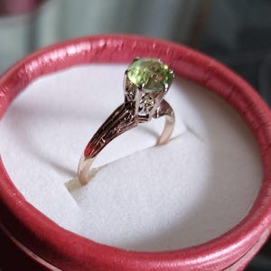 May include: A gold ring with a light green gemstone, set in a detailed filigree design. The ring is displayed inside a red ring box with a white interior, creating a striking contrast. The ring's band has intricate patterns.