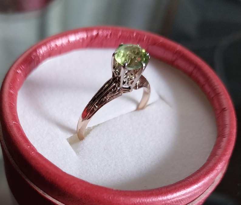May include: A gold ring with a light green gemstone, set in a detailed filigree design. The ring is displayed inside a red ring box with a white interior, creating a striking contrast. The ring's band has intricate patterns.