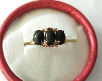 May include: A gold ring featuring three dark, oval-shaped stones. The stones are set in a decorative gold band with floral accents. The ring is presented in a red ring box with a white interior.