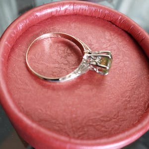 May include: A silver ring with a large, olive-green gemstone, set in a decorative, filigree-style mounting. The ring is displayed inside a round, red jewelry box, creating a striking contrast of colors and textures.