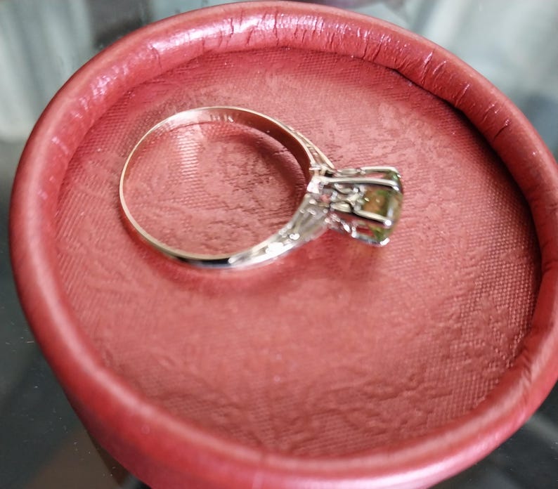 May include: A silver ring with a large, olive-green gemstone, set in a decorative, filigree-style mounting. The ring is displayed inside a round, red jewelry box, creating a striking contrast of colors and textures.