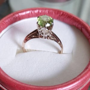 May include: A gold-toned ring with a light green gemstone, set in a detailed filigree design. The ring is presented in a red ring box with a white interior, showcasing the jewelry's elegance. The gemstone is round and faceted.